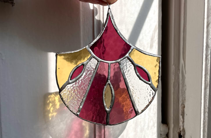 A hand holding a stained glass ornament with red, yellow, and clear sections, casting a colorful shadow on a white surface.