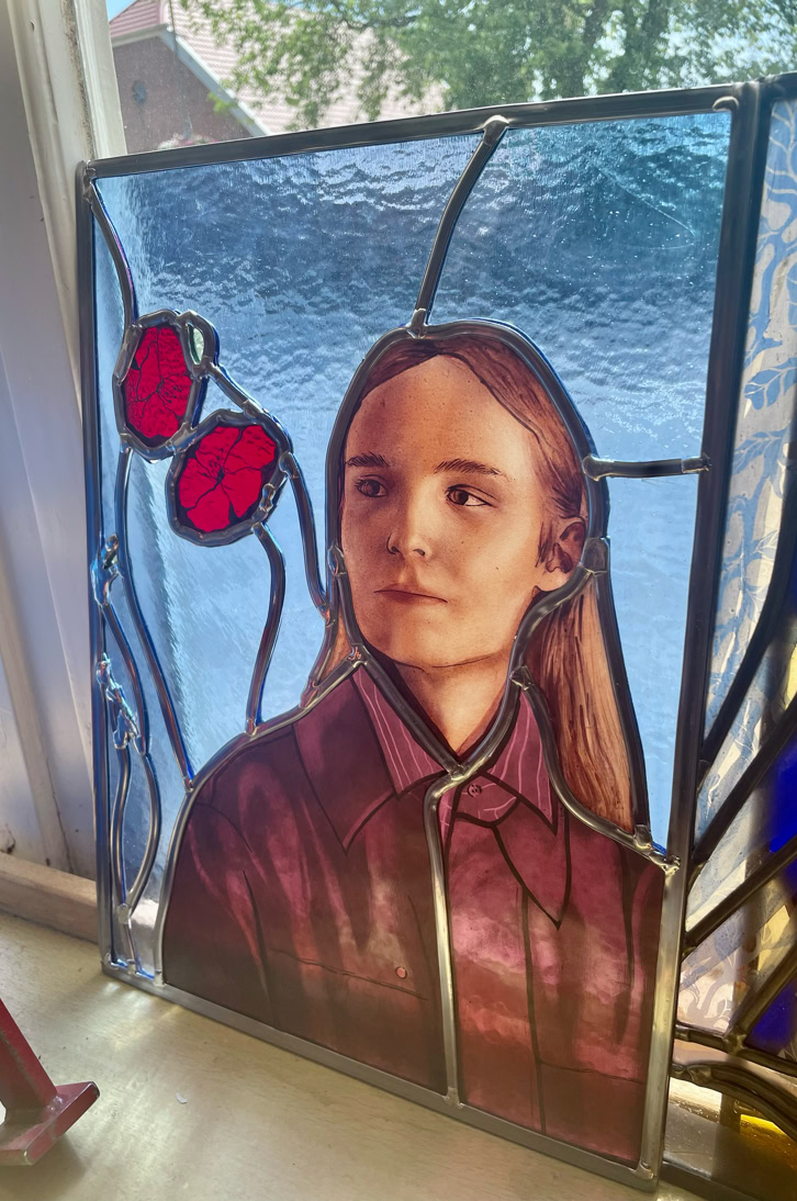 Stained glass artwork depicting a person with long hair and a collared shirt, with red flowers and a blue background.